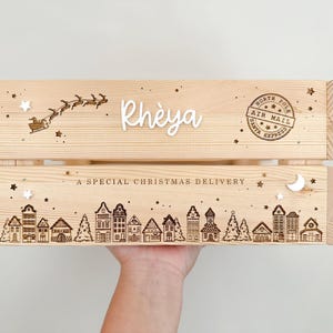May include: A light-colored wooden crate featuring laser-engraved details including a silhouette of Santa's sleigh, a North Pole Air Mail stamp, a row of houses, and the text "Rh&egrave;ya" and "A SPECIAL CHRISTMAS DELIVERY" with small star accents.