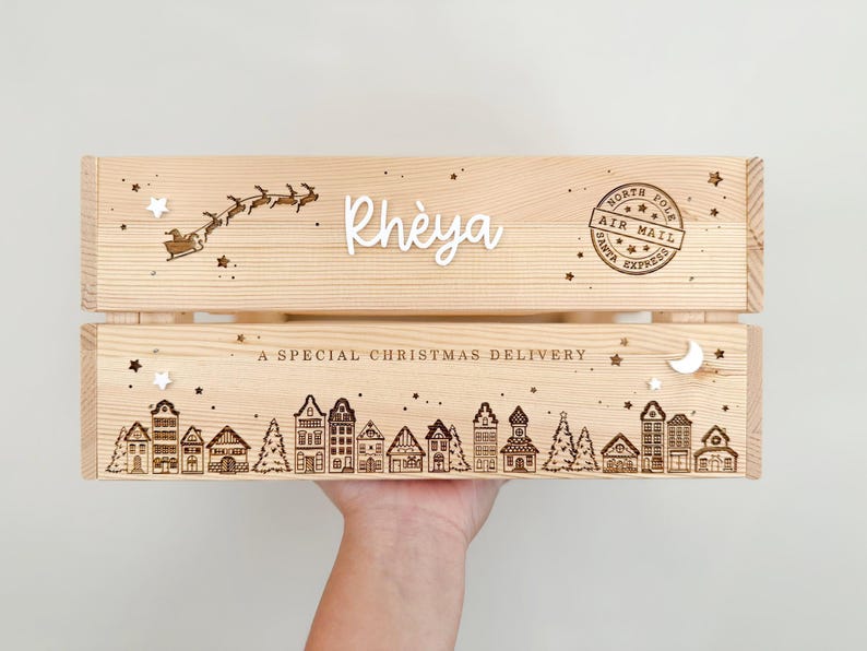 May include: A light-colored wooden crate featuring laser-engraved details including a silhouette of Santa's sleigh, a North Pole Air Mail stamp, a row of houses, and the text "Rh&egrave;ya" and "A SPECIAL CHRISTMAS DELIVERY" with small star accents.