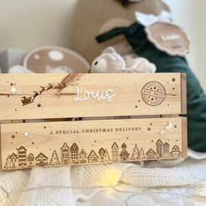 May include: A light-colored wooden crate featuring a laser-engraved North Pole air mail stamp, a silhouette of Santa in a sleigh, and a village scene. The name Louis is displayed in white script, with the text A SPECIAL CHRISTMAS DELIVERY.