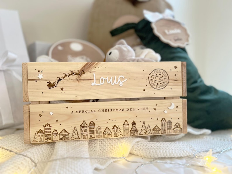 May include: A light-colored wooden crate featuring a laser-engraved North Pole air mail stamp, a silhouette of Santa in a sleigh, and a village scene. The name Louis is displayed in white script, with the text A SPECIAL CHRISTMAS DELIVERY.