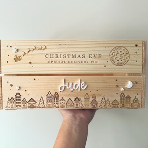May include: A light-colored wooden crate featuring engraved designs of a village skyline, stars, a crescent moon, and a Santa sleigh. The text reads CHRISTMAS EVE SPECIAL DELIVERY FOR, NORTH POLE AIR MAIL SANTA EXPRESS, and the name Jude in white script.