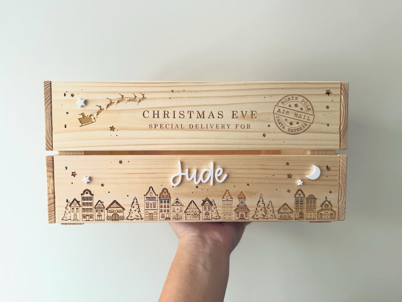 May include: A light-colored wooden crate featuring engraved designs of a village skyline, stars, a crescent moon, and a Santa sleigh. The text reads CHRISTMAS EVE SPECIAL DELIVERY FOR, NORTH POLE AIR MAIL SANTA EXPRESS, and the name Jude in white script.