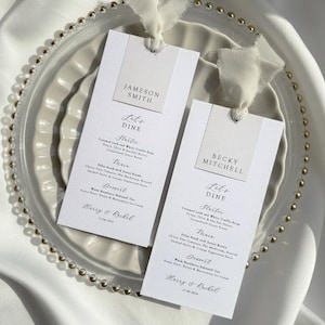 Puede incluir: Dos tarjetas de menú de boda con los nombres "Jameson Smith" y "Becky Mitchell". Las tarjetas blancas están atadas con cintas blancas y colocadas sobre un plato transparente con cuentas doradas. El fondo es blanco.