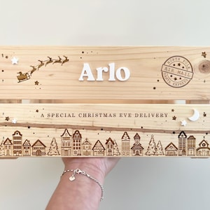 May include: A light-colored wooden crate featuring the name Arlo in white letters, a laser-engraved Santa sleigh, and a North Pole Air Mail stamp. The lower section displays a laser-engraved village scene with the text A SPECIAL CHRISTMAS EVE DELIVERY.