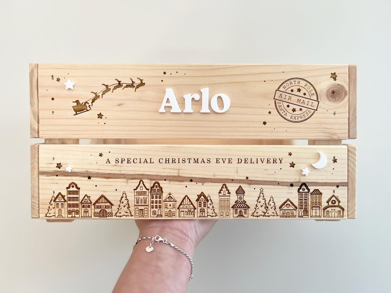 May include: A light-colored wooden crate featuring the name Arlo in white letters, a laser-engraved Santa sleigh, and a North Pole Air Mail stamp. The lower section displays a laser-engraved village scene with the text A SPECIAL CHRISTMAS EVE DELIVERY.