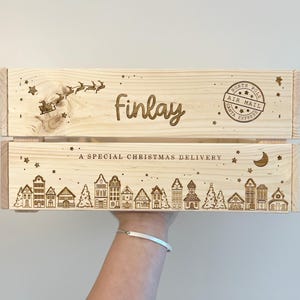 May include: A light-colored wooden crate featuring laser-engraved designs including a silhouette of Santa in a sleigh, the name "finlay," a "North Pole Air Mail" stamp, the text "A SPECIAL CHRISTMAS DELIVERY," and a row of village houses.