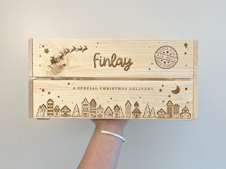 May include: A light-colored wooden crate featuring laser-engraved designs including a silhouette of Santa in a sleigh, the name "finlay," a "North Pole Air Mail" stamp, the text "A SPECIAL CHRISTMAS DELIVERY," and a row of village houses.