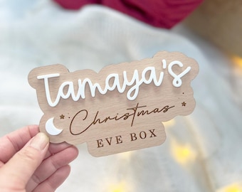 Pode incluir: Uma caixa de madeira para a véspera de Natal com o nome "Tamaya's" e o texto "Christmas Eve Box" em letras brancas. Um crescente lunar e estrelas também estão incluídos no design.