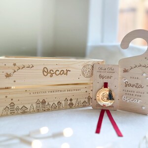 May include: A light-colored wooden crate engraved with the name Oscar, a sleigh, and a village scene, accompanied by a matching wooden door hanger reading Dear Santa Please Stop Here For Oscar and a gold-toned medal on a red ribbon.