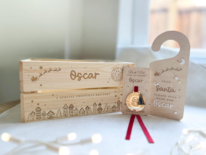 May include: A light-colored wooden crate engraved with the name Oscar, a sleigh, and a village scene, accompanied by a matching wooden door hanger reading Dear Santa Please Stop Here For Oscar and a gold-toned medal on a red ribbon.