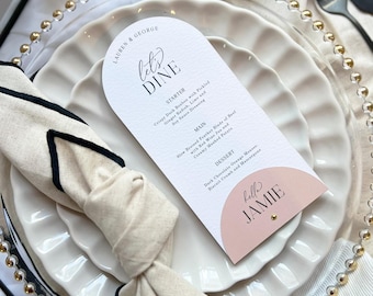 May include: An arched white paper menu card with black text reading Let's Dine, Lauren & George, and Jamie, featuring a pink semi-circle accent. The menu is placed on a white scalloped plate with a beige napkin featuring a black trim edge.
