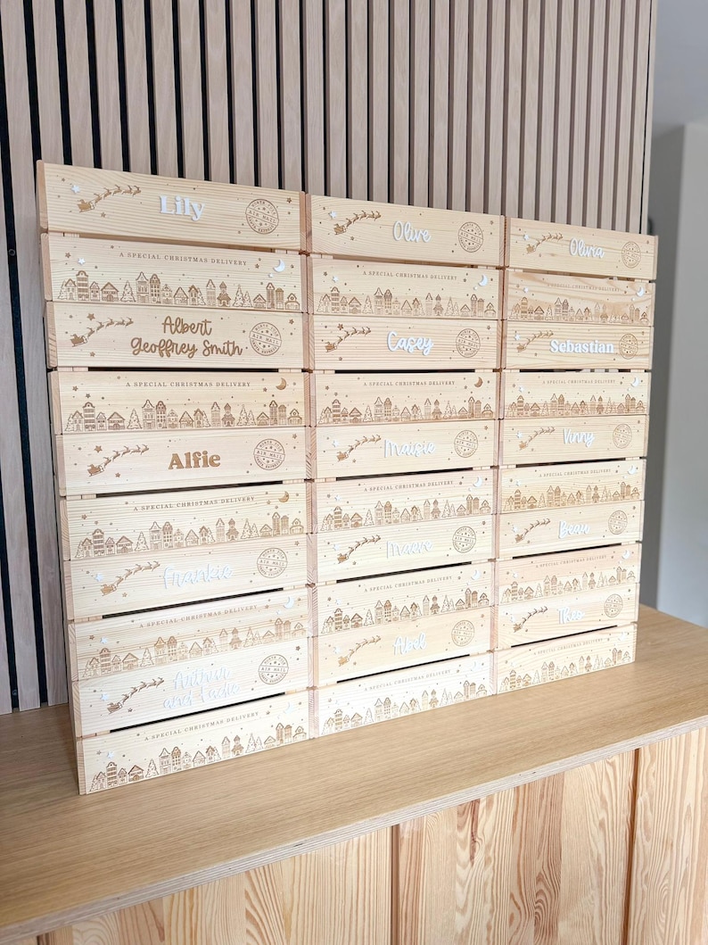 May include: A stack of light-colored wooden crates, each featuring a laser-engraved winter village scene, a silhouette of a sleigh with reindeer, and the text A SPECIAL CHRISTMAS DELIVERY. Each crate is personalized with a unique name.