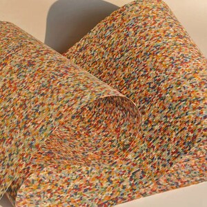 May include: Rolled paper with a colorful, speckled pattern. The design features a mix of red, blue, yellow, and green dots, creating a textured appearance. The paper is curled, revealing the pattern on the inside.