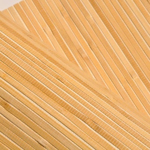 May include: Close-up of a bamboo mat in a natural, light brown hue. The mat is constructed from thin, parallel bamboo strips, providing a textured surface. The strips are sewn together with light-coloured thread, forming a chevron pattern.