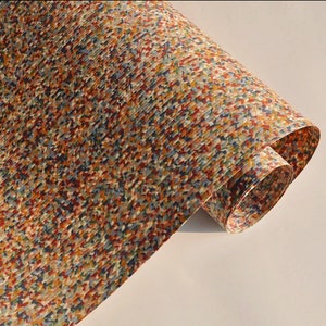 May include: A rolled sheet of paper with a colorful, speckled pattern. The design features small, irregular shapes in shades of red, blue, yellow, and brown, creating a textured appearance. The paper is slightly curled, revealing its underside.
