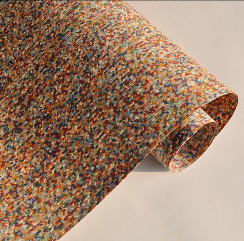 May include: A rolled sheet of paper with a colorful, speckled pattern. The design features small, irregular shapes in shades of red, blue, yellow, and brown, creating a textured appearance. The paper is slightly curled, revealing its underside.