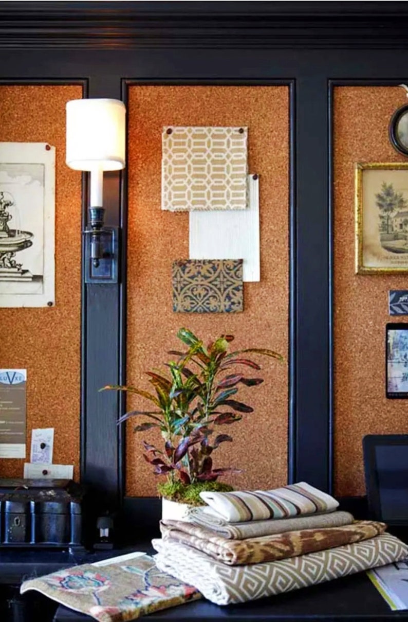 May include: A corkboard with various fabric swatches, a framed print, and a potted plant. The corkboard is mounted on a black wall. A stack of folded fabric is on a dark surface.