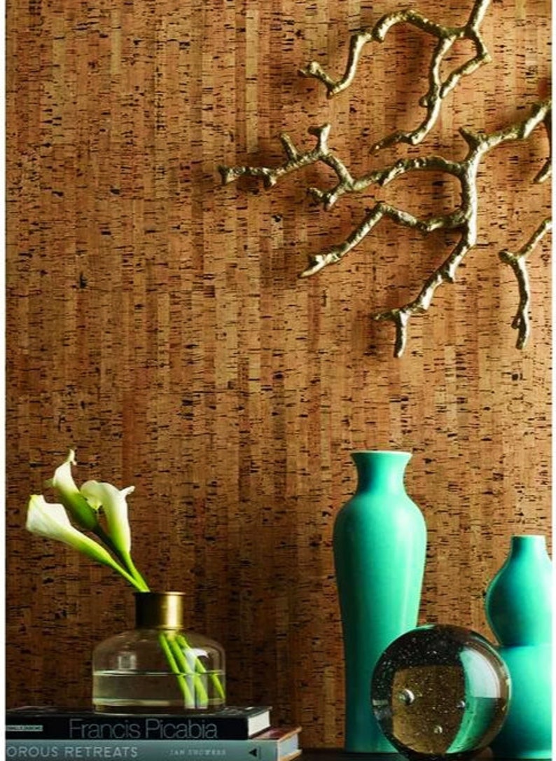 May include: A cork wall with a gold metal branch wall decor, a teal blue vase, a clear glass vase with white flowers, a clear glass orb, and a stack of books with the title "Francis Picabia" on the top book.
