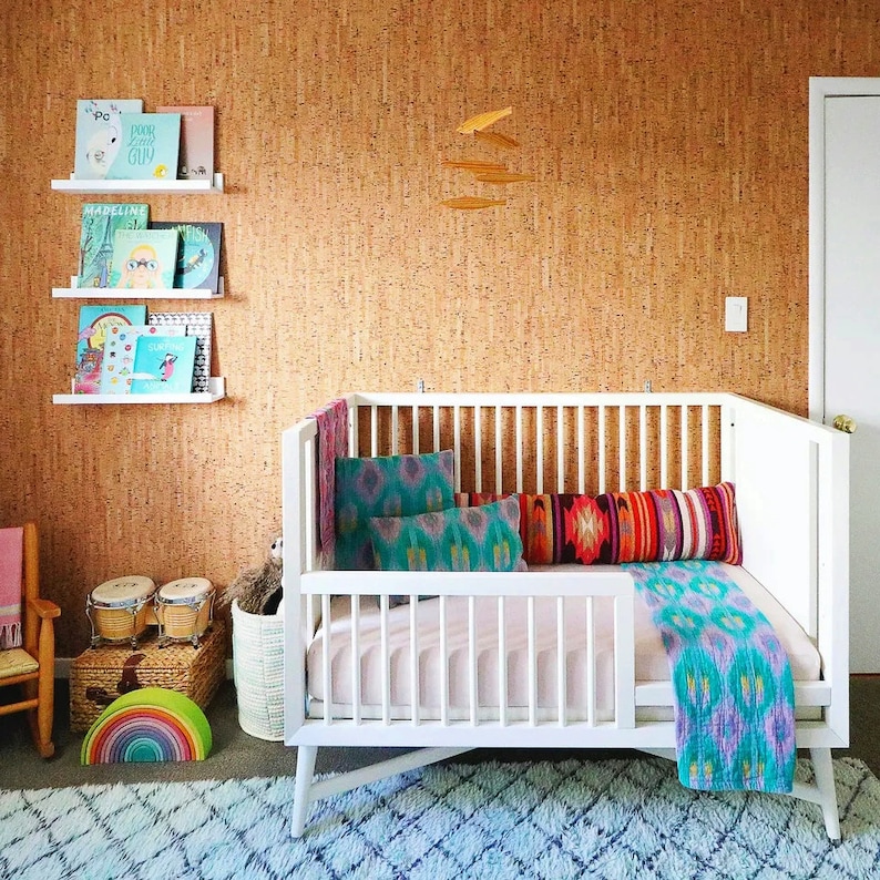 May include: A white cot with a pink mattress and a colourful blanket. The cot is against a cork wall with a mobile hanging above it. There are bookshelves on the wall with books on them. There are also some toys on the floor.