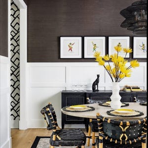 May include: A dining room with a black and white patterned wallpaper, a black and white patterned rug, a black table with a round top, and black chairs with woven seats. The table is set with yellow placemats and yellow plates. There are four framed pictures on the wall above the table.