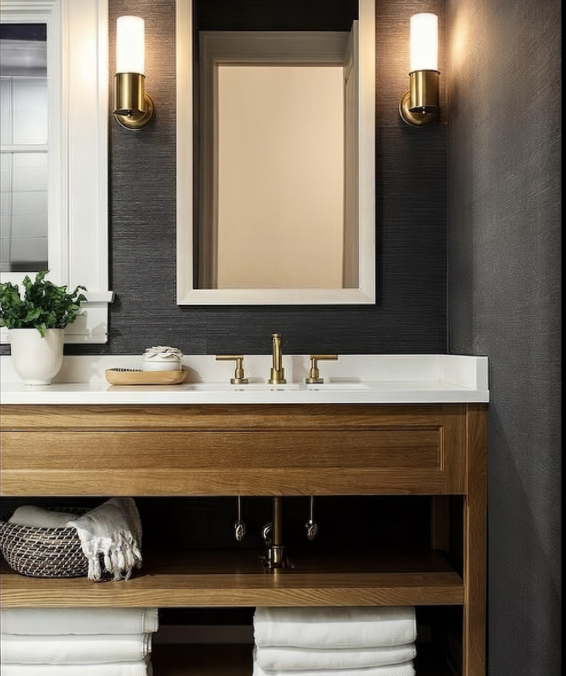 May include: A bathroom vanity with a white countertop and a wooden cabinet with open shelves. The vanity has a gold faucet and two gold wall sconces with frosted glass shades. The walls are dark gray and the shelves are filled with white towels.