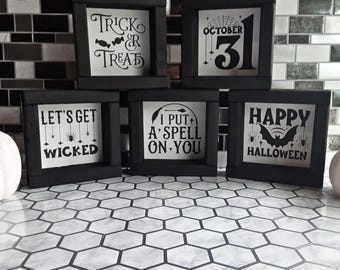 May include: Five black framed Halloween signs with white backgrounds. The signs display phrases like "Trick or Treat", "October 31", and "Happy Halloween". The signs are arranged on a patterned surface.