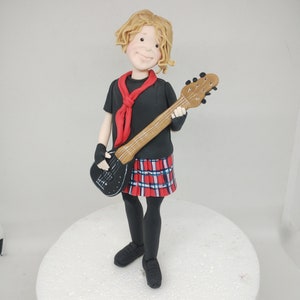 May include: A fondant figurine of a person with blonde hair wearing a black shirt, red scarf, and a plaid skirt. The person is holding a black and brown guitar.