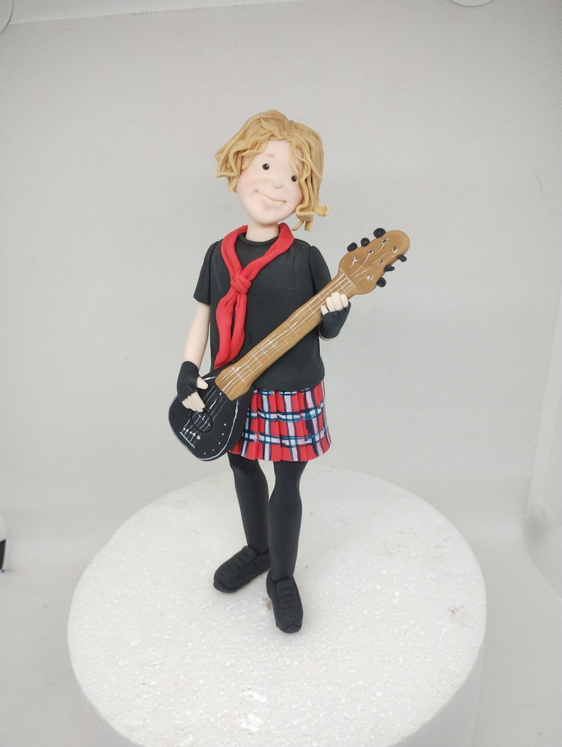 May include: A fondant figurine of a person with blonde hair wearing a black shirt, red scarf, and a plaid skirt. The person is holding a black and brown guitar.