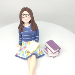 May include: A custom-made cake topper of a person sitting with a book and a stack of books. The person is wearing glasses, a blue and white striped shirt, a floral skirt, and has brown hair.