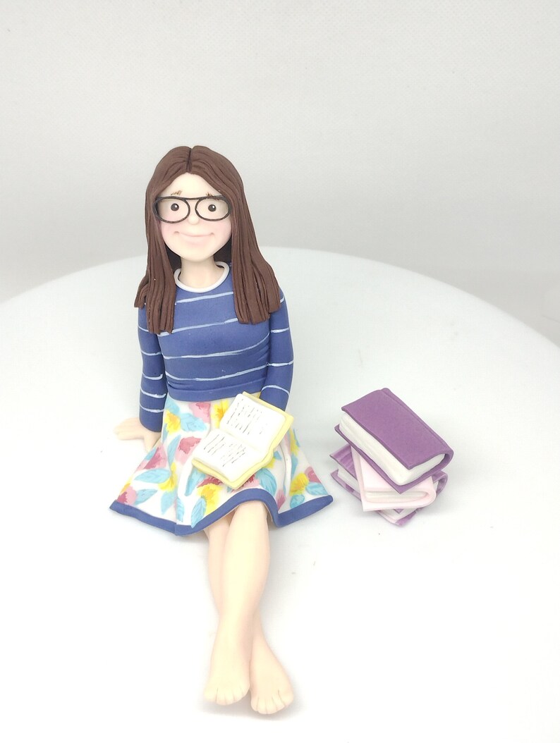 May include: A custom-made cake topper of a person sitting with a book and a stack of books. The person is wearing glasses, a blue and white striped shirt, a floral skirt, and has brown hair.