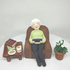 May include: A white-haired figurine of a person sitting in a brown armchair, holding a book. The person is wearing a green sweater and black pants. A small table with a cup, cookies, and books is to the left of the chair. A potted plant with white flowers is to the right of the chair.