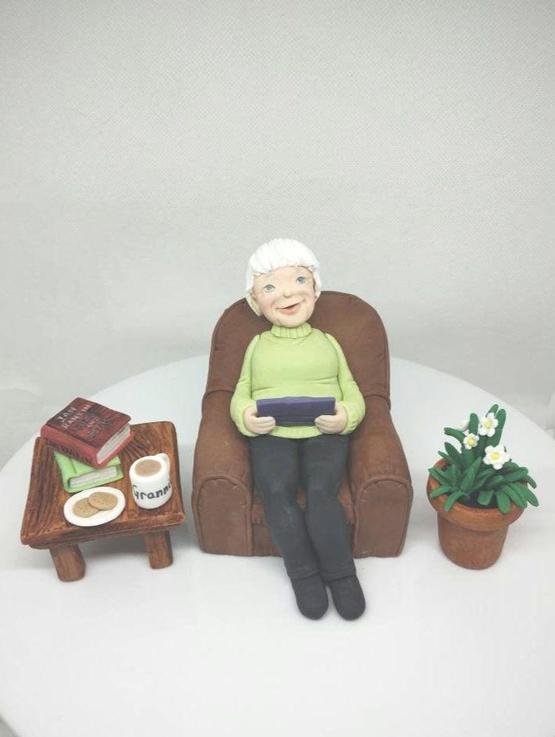 May include: A white-haired figurine of a person sitting in a brown armchair, holding a book. The person is wearing a green sweater and black pants. A small table with a cup, cookies, and books is to the left of the chair. A potted plant with white flowers is to the right of the chair.