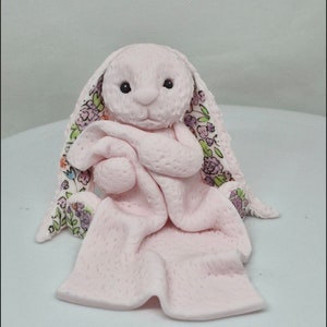 Op de afbeelding: Een lichtroze gebeeldhouwd konijnenfiguurtje met een getextureerd oppervlak, dat een bijpassend roze dekentje vasthoudt. Het konijn heeft lange oren met een bloemenpatroon aan de binnenkant, met paarse, groene en oranje bloemen op een witte achtergrond.