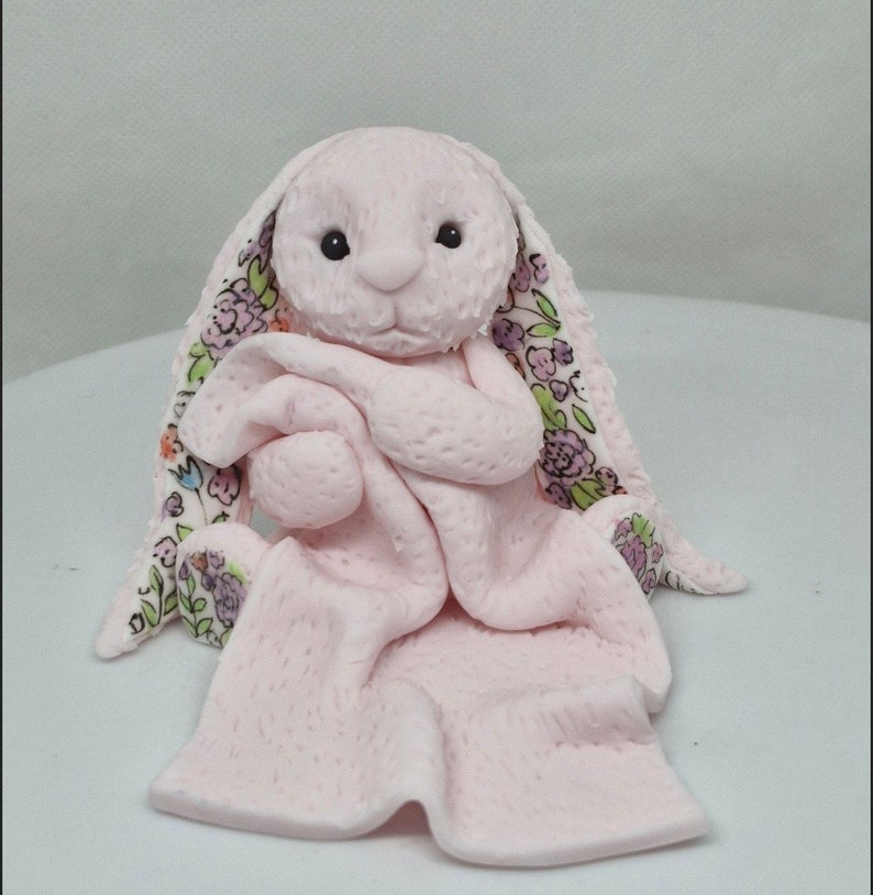 Op de afbeelding: Een lichtroze gebeeldhouwd konijnenfiguurtje met een getextureerd oppervlak, dat een bijpassend roze dekentje vasthoudt. Het konijn heeft lange oren met een bloemenpatroon aan de binnenkant, met paarse, groene en oranje bloemen op een witte achtergrond.