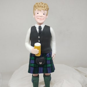 May include: A figurine of a person wearing a black vest, white shirt, and a green, blue, and black kilt. The person is holding a yellow can in their right hand. The figurine is standing on a white surface.