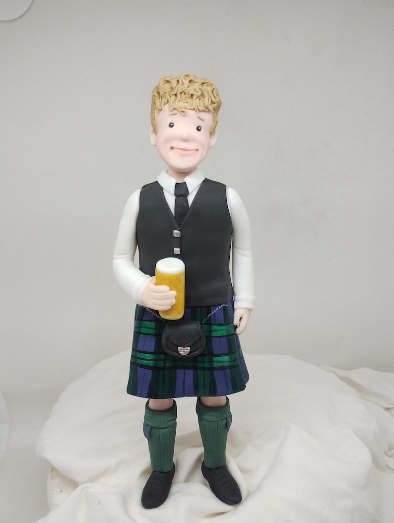 May include: A figurine of a person wearing a black vest, white shirt, and a green, blue, and black kilt. The person is holding a yellow can in their right hand. The figurine is standing on a white surface.