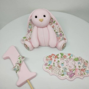 Op de afbeelding: Een lichtroze fondant konijnfiguurtje met bloemmotief aan de binnenkant van de oren en voeten, een taarttopper in de vorm van het cijfer &eacute;&eacute;n met een bloemstrik, en een geschulpt bordje met de naam ROSE in lichtroze letters op een bloemenachtergrond.