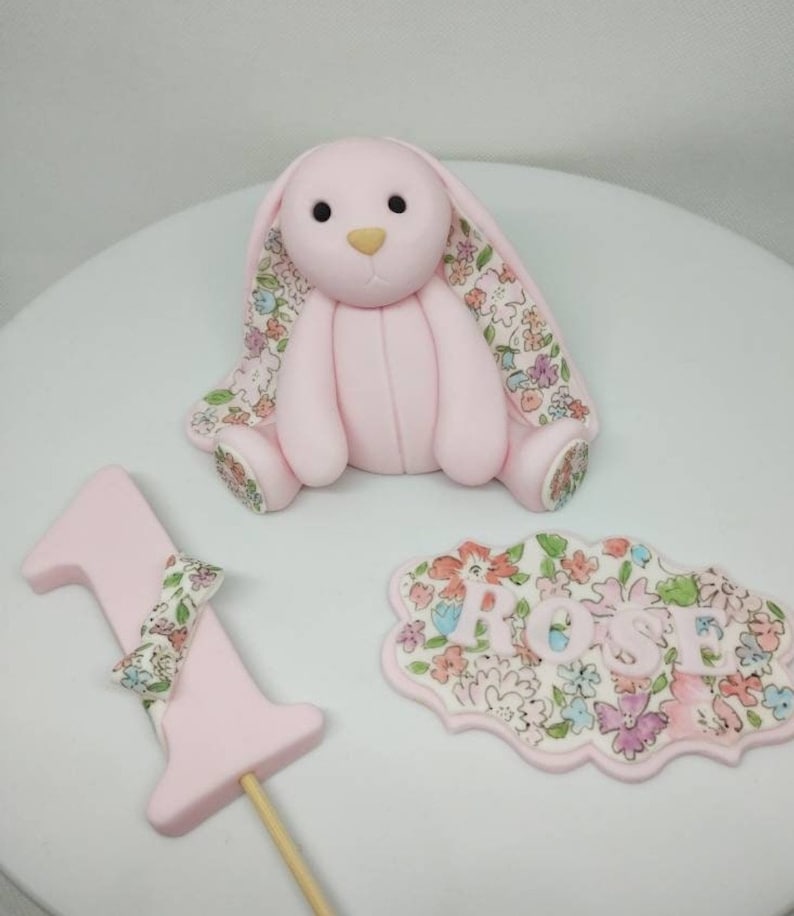 Op de afbeelding: Een lichtroze fondant konijnfiguurtje met bloemmotief aan de binnenkant van de oren en voeten, een taarttopper in de vorm van het cijfer &eacute;&eacute;n met een bloemstrik, en een geschulpt bordje met de naam ROSE in lichtroze letters op een bloemenachtergrond.