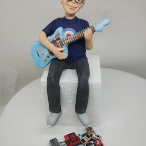May include: A blue and white guitar with a seated figurine of a person wearing glasses and a blue shirt with a target symbol. The figurine is holding the guitar. There are several small, colorful, rectangular objects in front of the figurine.