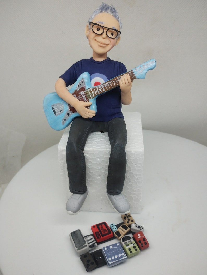 May include: A blue and white guitar with a seated figurine of a person wearing glasses and a blue shirt with a target symbol. The figurine is holding the guitar. There are several small, colorful, rectangular objects in front of the figurine.
