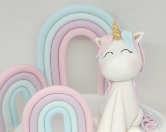 May include: A white unicorn cake topper with a gold horn and a pink and blue mane. The unicorn is sitting in front of a pastel rainbow.