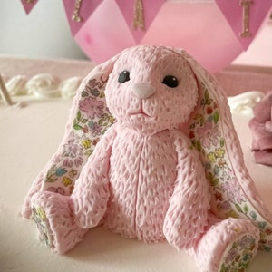 Op de afbeelding: Een roze konijnenfiguur met een getextureerd, vachtachtig oppervlak, zwarte ogen en een witte neus. De binnenkant van de oren en de voetzolen zijn voorzien van een kleurrijk bloemenpatroon met kleine blauwe, gele, roze en groene bloemetjes.