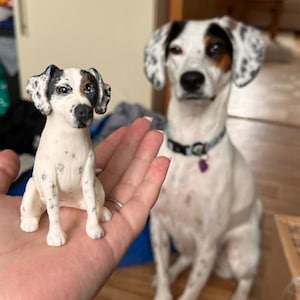 May include: A small, white dog figurine with black and brown markings, held in a hand. The figurine is a miniature replica of a dog with similar markings, sitting in the background. The dog in the background is white with black and brown spots.
