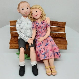 May include: A wedding cake topper featuring a couple sitting on a wooden bench. The woman is wearing a pink floral dress and the man is wearing a plaid shirt and shorts.