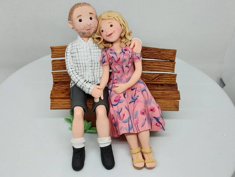 May include: A wedding cake topper featuring a couple sitting on a wooden bench. The woman is wearing a pink floral dress and the man is wearing a plaid shirt and shorts.