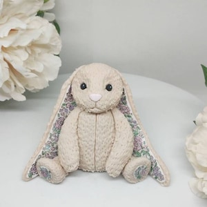 Op de afbeelding: Een beige pluche konijnenspeeltje met een textuur die op vacht lijkt, zwarte kraaloogjes en een lichtroze neus. De lange, slappe oren zijn gevoerd met een witte stof met een bloemenpatroon van kleine paarse, roze en groene bloemetjes.