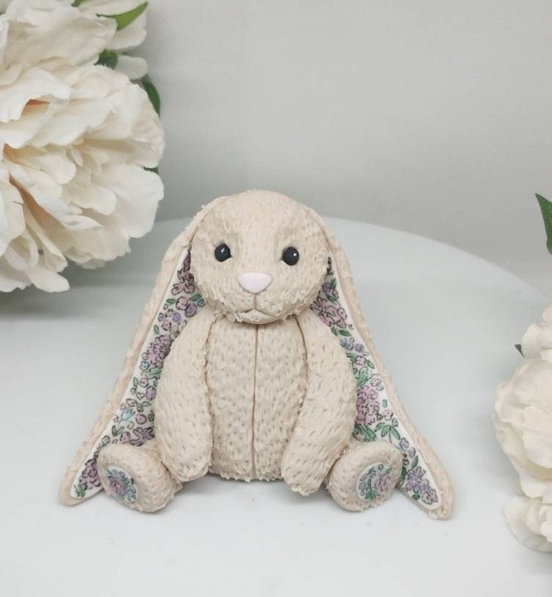 Op de afbeelding: Een beige pluche konijnenspeeltje met een textuur die op vacht lijkt, zwarte kraaloogjes en een lichtroze neus. De lange, slappe oren zijn gevoerd met een witte stof met een bloemenpatroon van kleine paarse, roze en groene bloemetjes.
