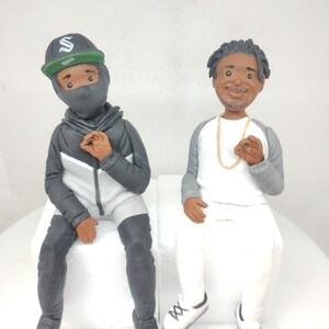 May include: Two figurines of people sitting on white blocks. The person on the left is wearing a black hooded jacket, black pants, a black mask, and a green and black baseball cap with a white letter "S" on it. The person on the right is wearing a white shirt, gray pants, and white sneakers with black laces. They are both smiling.