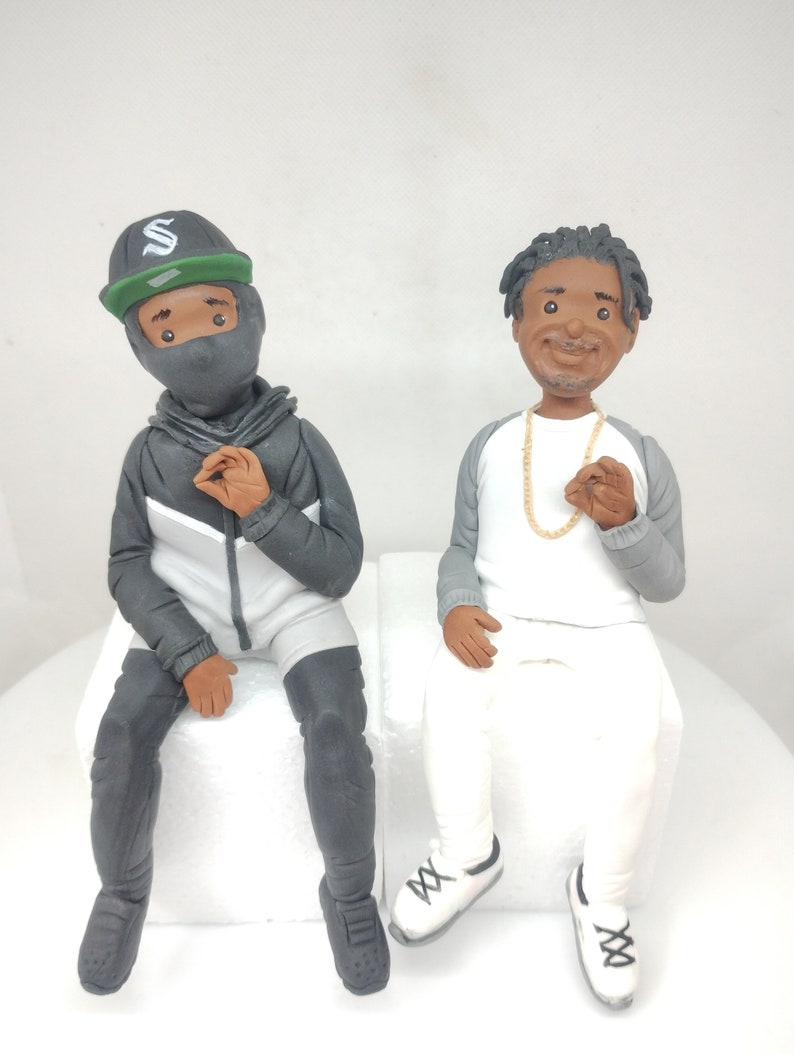 May include: Two figurines of people sitting on white blocks. The person on the left is wearing a black hooded jacket, black pants, a black mask, and a green and black baseball cap with a white letter "S" on it. The person on the right is wearing a white shirt, gray pants, and white sneakers with black laces. They are both smiling.