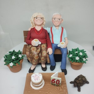 May include: A 60th anniversary cake with a couple sitting on a bench, holding cups of tea. The cake is decorated with a small cake, a card with the number 60, and a small turtle.